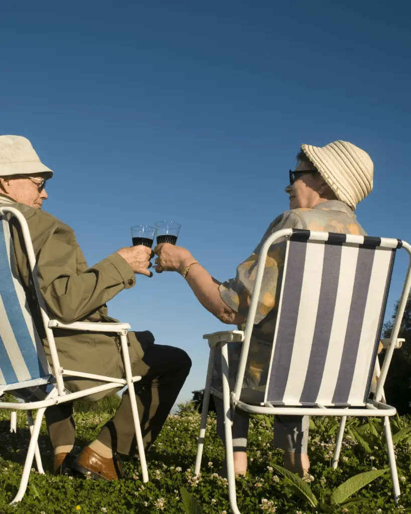 Starejši par se drži za roke in nazdravlja z vinom na stolih pod modrim nebom
